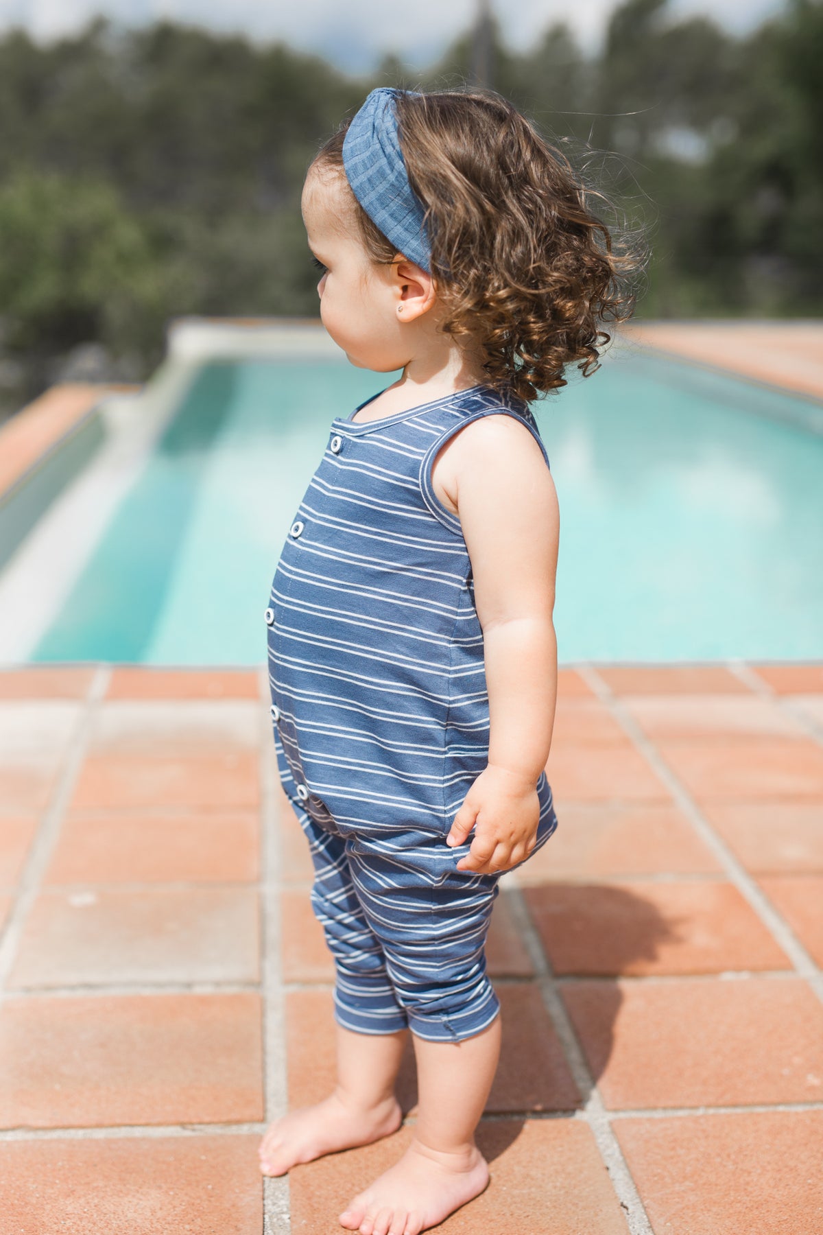 Little girl standing by a pool barefoot wearing blue and white striped sleeveless overalls by 1+ In The Family.